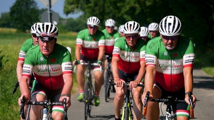 Eine Gruppe Fahrradfahrer, die mit grün-weiß-roten Trikots und weißen Helmen durch die Landschaft fährt.