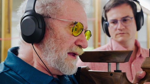 Jürgen Klamann beim Schießen. Er trägt eine Brille mit gelben Gläsern. Neben ihm steht sein Trainer, beide haben Kopfhörer auf.