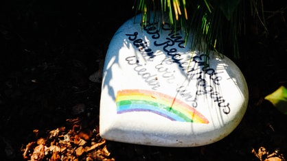Ein kleines Herz mit Aufschrift und einem Regenbogen liegt auf einem Grab
