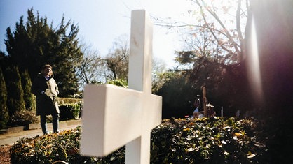 Auf einem Friedhof steht ein weißes Kreuz aus Holz, im Hintergrund steht Susanne Starkmuth vor einem Grab
