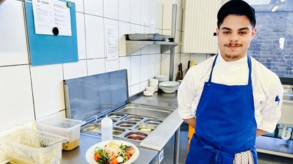 Salvatore Longobardo steht gekleidet in Schürze und Kappe neben einer Küchenzeile.
