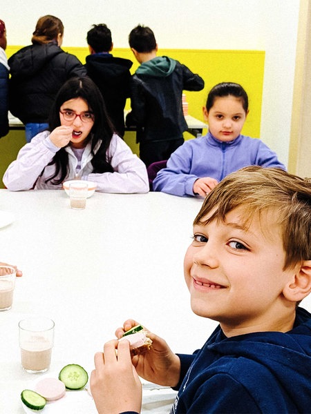 Einige Kinder sitzen an einem großen grauen Tisch. Einige essen ihr Frühstück.