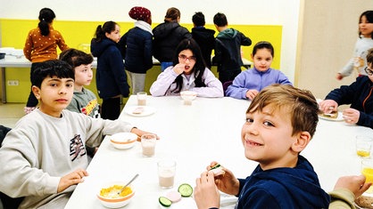 Einige Kinder sitzen an einem großen grauen Tisch. Einige essen ihr Frühstück.
