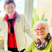 Die zwei älteren Frauen Magret Pöppel und Anneliese König sitzen an einem Tisch und lächeln in die Kamera. Buchspaziererin Anette Fath steht zwischen ihnen und legt ihren Arm um deren Schultern.