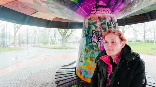Susan sitzt in einem Park auf einer überdachten Bank