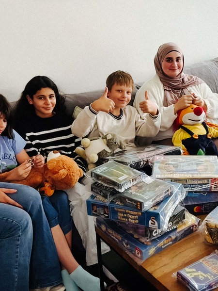 Eine Gruppe von Kindern und einer Frau sitzt auf einer Couch. Vor ihnen ist ein Tisch mit verpackten Spielen.