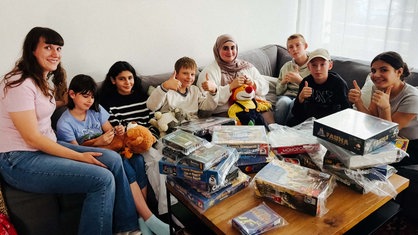 Eine Gruppe von Kindern und einer Frau sitzt auf einer Couch. Vor ihnen ist ein Tisch mit verpackten Spielen.