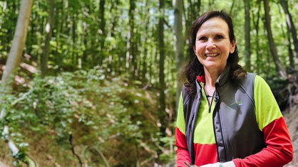 Claudia Herzog steht in einem Waldabschnitt des Siebengebirges. Sie trägt eine Jacke des Verschönerungsvereins und lächelt in die Kamera.