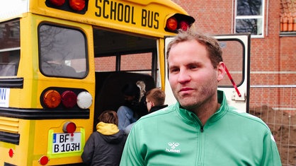 Der Lehrer Daniel Peitz sthet in einer grünen Jacke vor einem gelben Schulbus