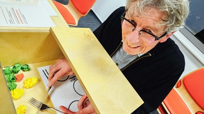 Michael Philipps, graue Haare, schwarze Brille, sitzt vor einem Holzkasten, in dem er über einen Spiegel versucht, kleine Papierkugeln mit Messer und Gabel aufzunehmen.