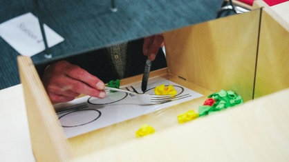 Ein Spiegel in einem Holzkasten zeigt spiegelverkehrt, wie ein Mann Messer und Gabel in den Händen hält und versucht, damit Papierkugeln aufzunehmen.