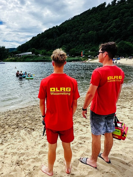 Henning Thielemeyer und Philipp Fögen stehen am Strand vom Godelheimer See und blicken auf den See, auf ihren roten T-Shirts steht auf dem Rücken "DLRG Wasserrettung"