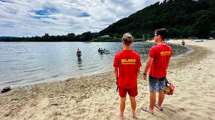 Henning Thielemeyer und Philipp Fögen stehen am Strand vom Godelheimer See und blicken auf den See, auf ihren roten T-Shirts steht auf dem Rücken "DLRG Wasserrettung"