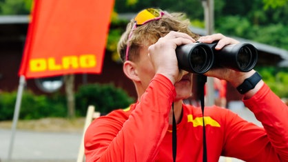 Henning Thielemeyer sitzt in einem Strandstuhl und hält ein Fernglas vor die Augen, im Hintergrund weht eine rote Flagge mit den gelben Buchstaben "DLRG"