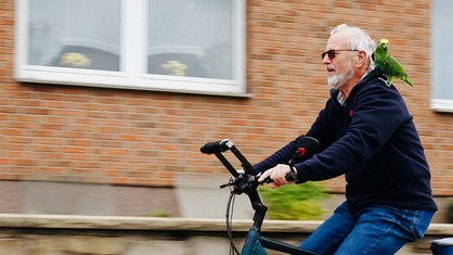 Wolfgang Busse fährt mit Sonnenbrille Fahrrad. Auf seiner linken Schulter sitzt ein Papagei.
