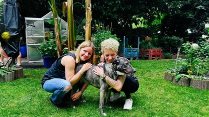Zwei Frauen, Nadin Duberg und Susi Schmücker, hocken in einem Garten und umarmen ihre grau-weiße Hündin Wally.