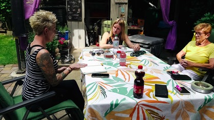Nadin Duberg und Susi Schmücker sitzen an einem Tisch, jeweils einen Kalender vor ihnen auf dem Tisch liegend, rechts daneben sitzt Susis Mutter Hanne, auf dem Tisch liegen Handys und stehen Flaschen.