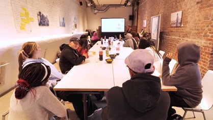 Jugendliche mit dem Nordstern-Stipendium sitzen in einem Raum mit geklinkerten Wänden an einem langen Tisch und gucken auf eine große Leinwand