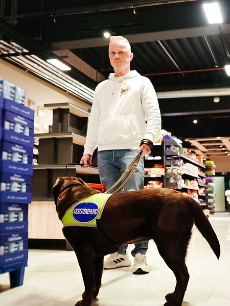Ein Mann steht im Supermarkt. An der Leine hält er einen braunen Labrador.