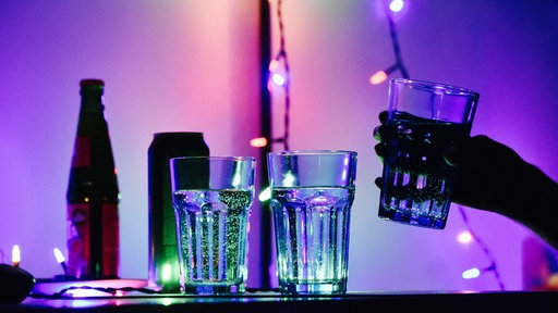 Drei Gläser gefüllt mit einer durchsichtigen Flüssigkeit stehen auf einem Tisch. Dahinter eine Flasche und eine Dose. Die Beleuchtung ist dunkel.