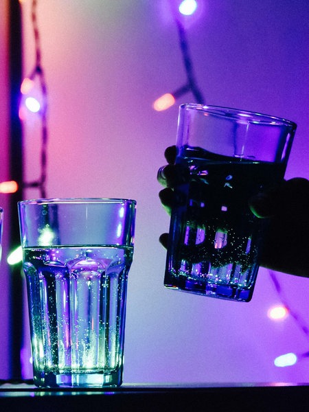 Drei Gläser gefüllt mit einer durchsichtigen Flüssigkeit stehen auf einem Tisch. Dahinter eine Flasche und eine Dose. Die Beleuchtung ist dunkel.
