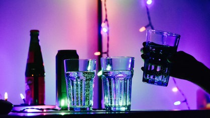 Drei Gläser gefüllt mit einer durchsichtigen Flüssigkeit stehen auf einem Tisch. Dahinter eine Flasche und eine Dose. Die Beleuchtung ist dunkel.