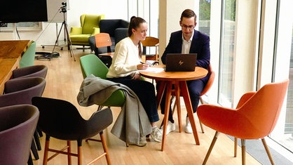 Tabea Roßhoff, eine junge Frau, und ein Mann, Ole Schley, sitzen auf gepolsterten bunten Stühlen hinter einem runden Tisch und schauen konzentriert auf einen Bildschirm.