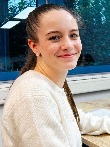 Tabea Roßhoff, eine junge brünette Frau im weißen Strickpullover, sitzt an einem Tisch vor ihrem Laptop, neben ihr stehen Gesetzbücher. Sie schaut freundlich in die Kamera.