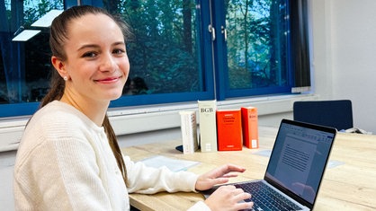 Tabea Roßhoff, eine junge brünette Frau im weißen Strickpullover, sitzt an einem Tisch vor ihrem Laptop, neben ihr stehen Gesetzbücher. Sie schaut freundlich in die Kamera.