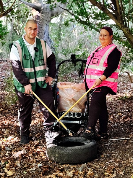 Marvin Rathmann in grüner Warnweste und Clarissa Rathmann in pinker Warnweste stehen in einem Wald und lächeln in die Kamera. In der Hand halten sie Zangen, in ihrer Mitte steht ein Kinderwagen mit einer Mülltüte.