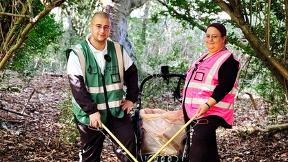 Marvin Rathmann in grüner Warnweste und Clarissa Rathmann in pinker Warnweste stehen in einem Wald und lächeln in die Kamera. In der Hand halten sie Zangen, in ihrer Mitte steht ein Kinderwagen mit einer Mülltüte.
