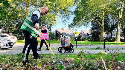 Marvin Rathmann in grüner Warnweste hebt mit einer Zange eine Plastiktüte auf, im Hintergrund läuft Clarissa Rathmann in pinker Warnweste. Neben ihr steht ein Kinderwagen mit Mülltüten.