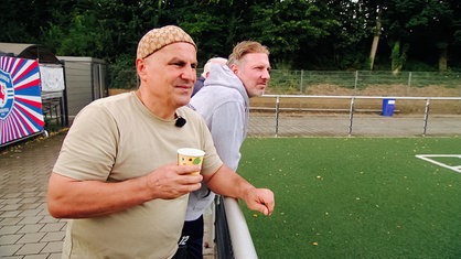 Ehrenamtler Suat Amaҫ steht mit einem Becher in der Hand neben Spielerinnen-Vater David Meyer am Spielfeldrand.