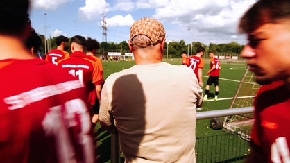 Suat Amaҫ von hinten. Er steht an einem Fußballfeld, um ihm herum laufen Fußballspieler.