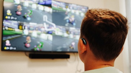 Ein Kind spielt Mario Kart. Man sieht nur den Hinterkopf vom Kind und im Hintergrund ist ein Bildschirm mit dem Spiel.