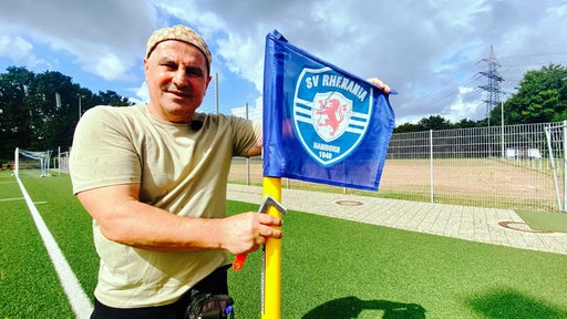 Suat Amaç, ein trainierter Mann mittleren Alters mit einer braunen Kappe und einem beigen T-Shirt hält eine blaue Eckfahne auf dem Fußballplatz.