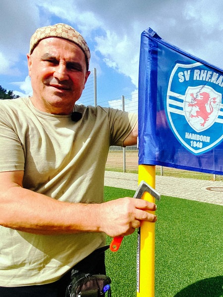Suat Amaç, ein trainierter Mann mittleren Alters mit einer braunen Kappe und einem beigen T-Shirt hält eine blaue Eckfahne auf dem Fußballplatz.
