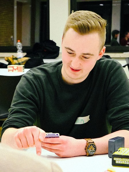 Jan Berkemeyer, ein junger blonder Mann im grünen Pullover, sitzt hinter einem weißen Tisch und schaut auf sein Smartphone, das er in den Händen hält.