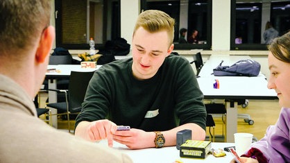 Jan Berkemeyer, ein junger blonder Mann im grünen Pullover, sitzt hinter einem weißen Tisch und schaut auf sein Smartphone, das er in den Händen hält.