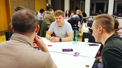 In einem hellen Raum sitzen zehn junge Menschen an zwei weißen Tischen verteilt und spielen gemeinsam Gesellschaftsspiele.