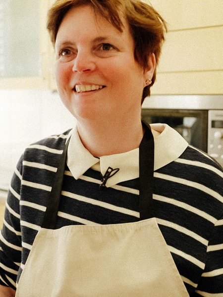 Eine Frau mit kurzen hellbraunen Haaren lächelt nach vorne in die Kamera, sie trägt ein blau-weiß-gestreiftes Hemd, darüber eine weiße Schürze