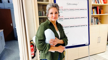 Susanna Jochims steht mit einem herzförmigen Kissen unter dem Arm vor einem Flipchart mit Erklärungen zum Nähen. Sie schaut in die Kamera.