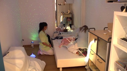 Ein kleines Mädchen liegt auf einer Matratze und hält kurze Lichterketten. Zwei Pflegerinnen sind bei ihr.