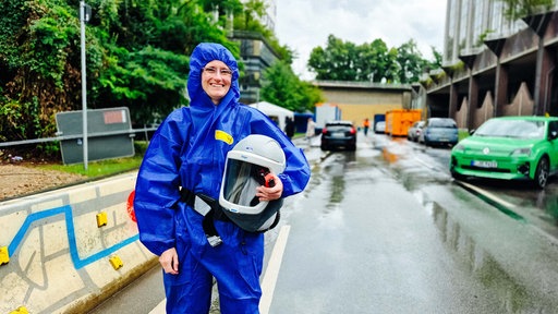 Eine Frau steht in Schutzkleidung auf der Straße und lächelt in die Kamera.