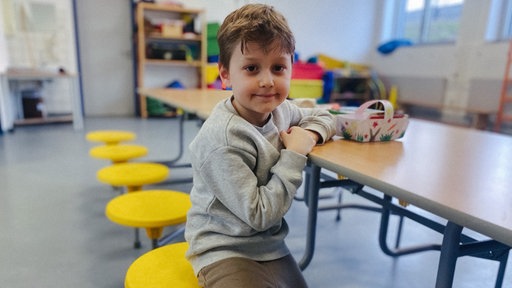 Der sechsjährige Loukas sitzt an einem Tisch und lächelt in die Kamera.
