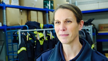 Katharina Kluge im Portrait in einer Lagerhalle. Im Hintergrund hängen Feuerwehrschutzanzüge an einer Kleiderstange.