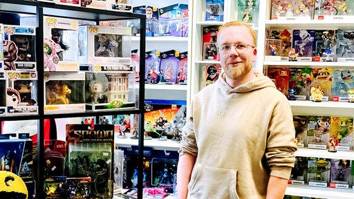 Ein Mann mit Kapuzenpulli steht neben einer Vitrine mit Actionfiguren, hinter ihm weitere Regale: