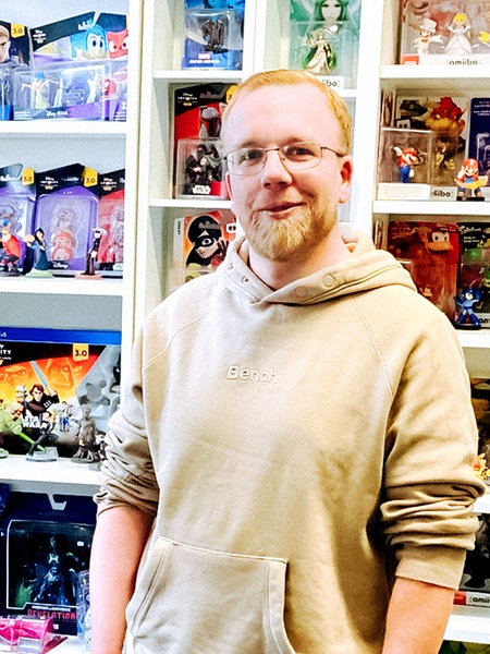 Ein Mann mit Kapuzenpulli steht neben einer Vitrine mit Actionfiguren, hinter ihm weitere Regale: