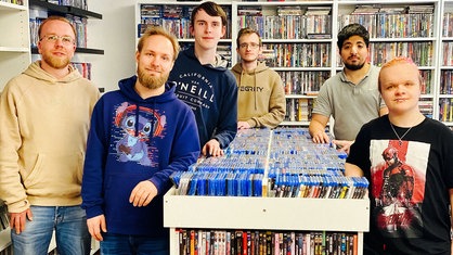 Sechs Männer stehen vor Regalen mit DVDs und blicken in die Kamera