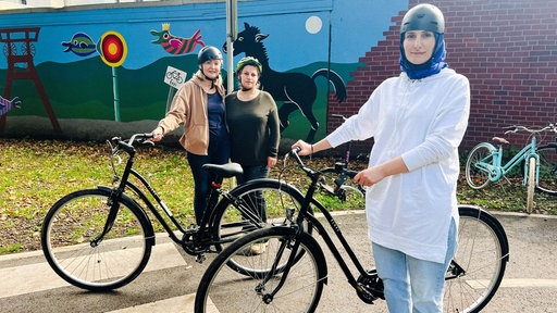 Drei Frauen mit Helm stehen neben zwei Fahrrädern auf der Straße.